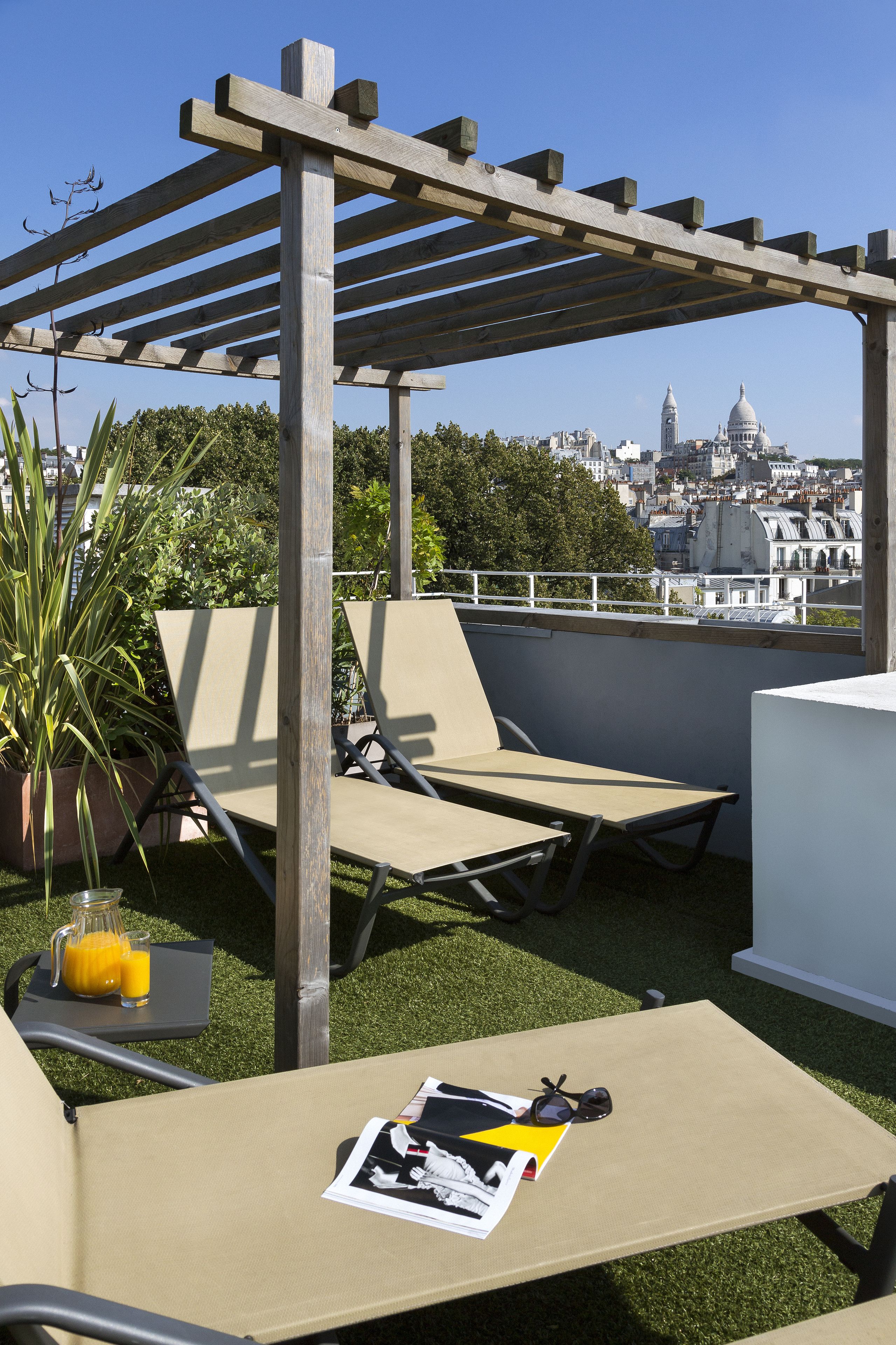 A rooftop terrace with two lounge chairs, a table with a drink and a magazine, and a pergola structure.