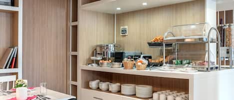 Una barra de desayuno equipada con cafetera, tostadora y diversos alimentos. Se observan estantes con platos, tazones y tazas.