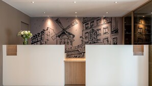 A reception area with a modern design, featuring a large mural of a windmill and buildings, a wooden reception desk, and a vase with flowers. - Citadines Montmartre Paris (Paris)