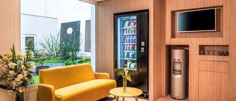 A modern hotel room with a yellow sofa, a small round table, a refrigerator, a water dispenser, and a TV mounted on the wall.