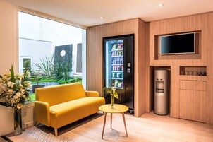 Quarto de hotel moderno com um sofá amarelo, uma pequena mesa redonda, um frigorífico, um dispensador de água e uma televisão montada na parede.