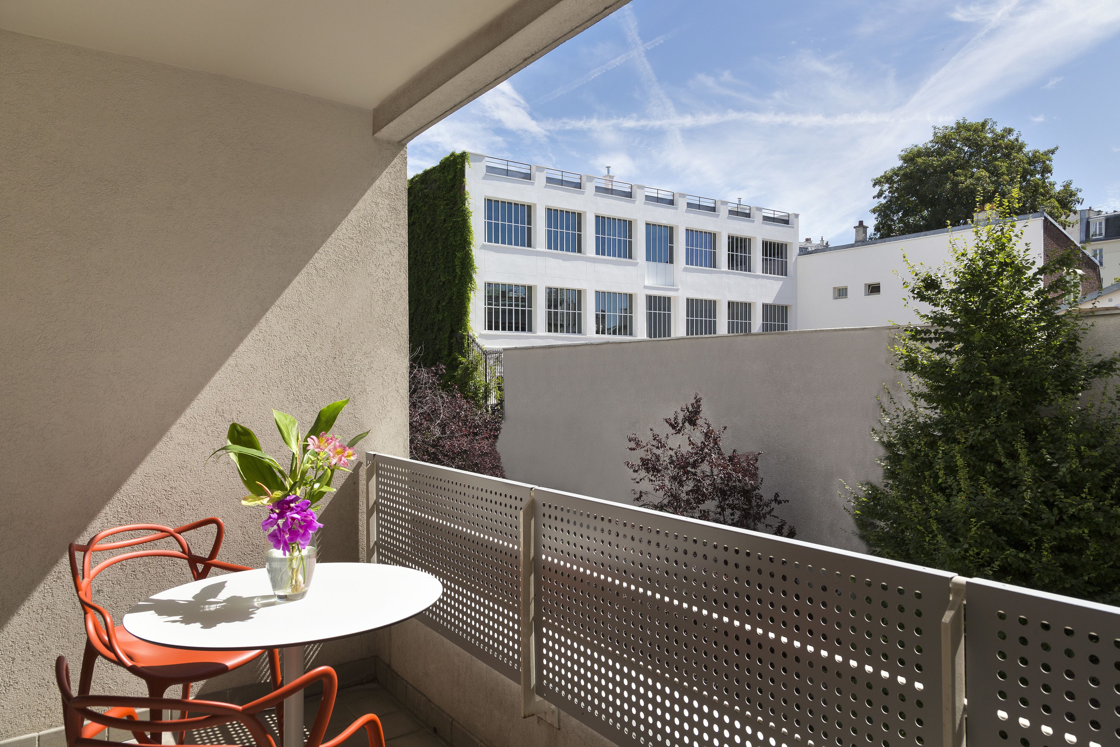 A balcony with a round table and two chairs, a potted plant, and a view of a building with multiple windows and a tree.