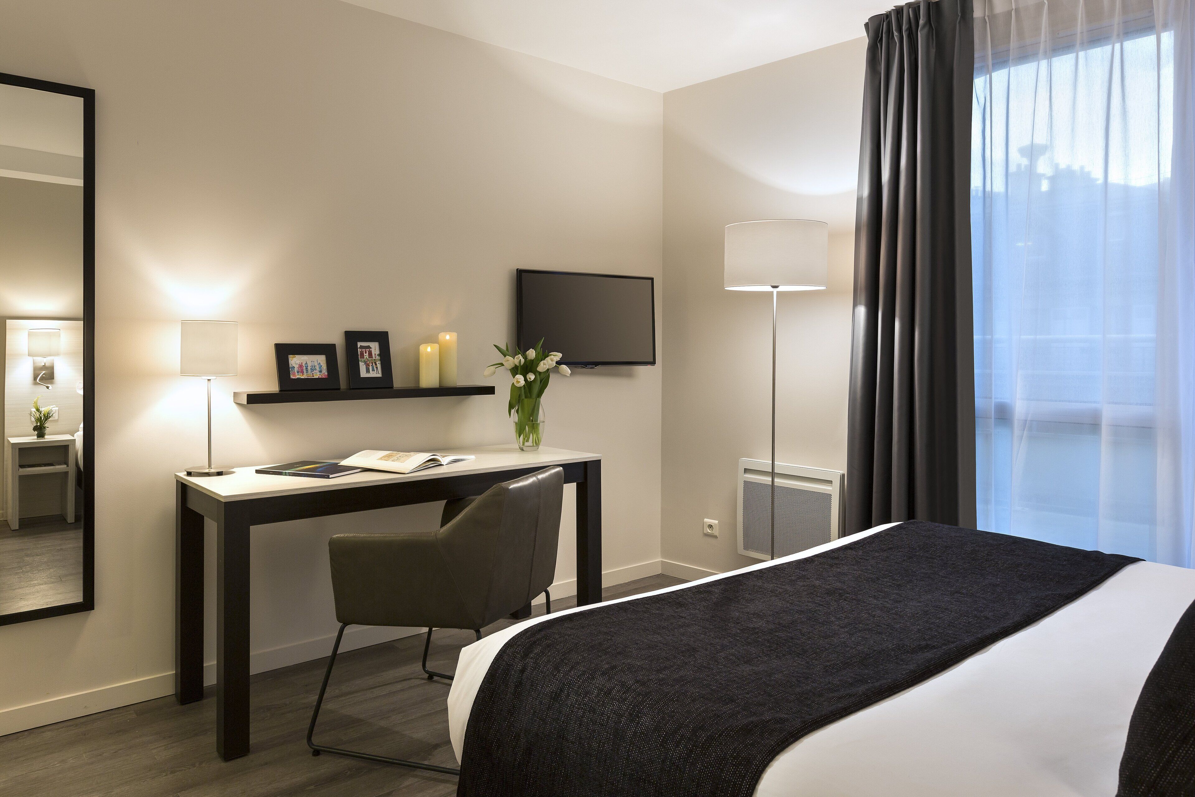 A hotel room with a bed, a desk with a chair, a TV, a lamp, and a window with curtains.