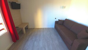 Ein Zimmer mit einem braunen Sofa, einer kleinen Holzbank, einem an der Wand befestigten Flachbildfernseher und einem Fenster mit roten Vorhängen.