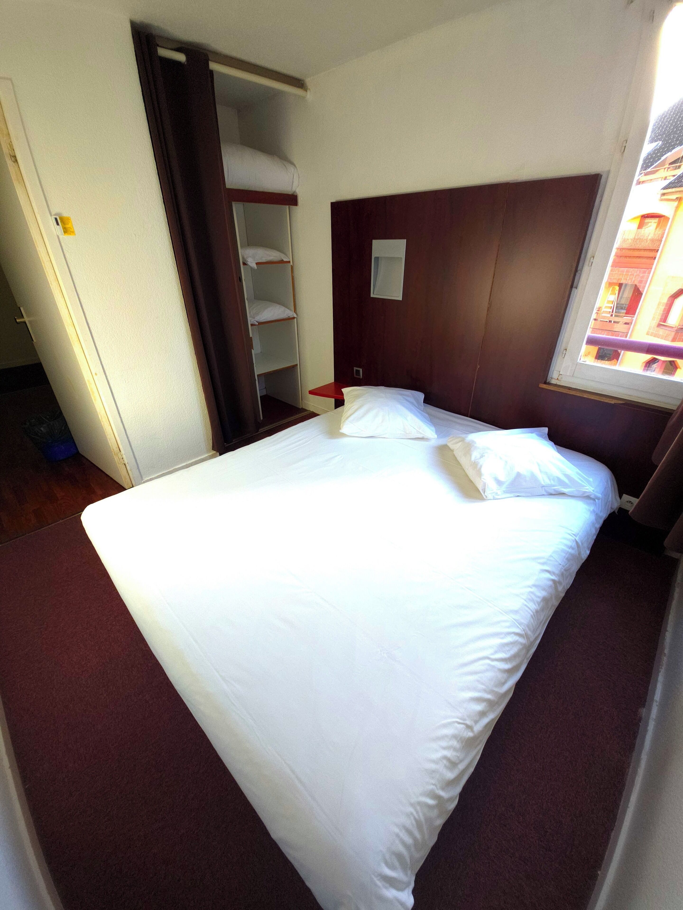 A hotel room with a bed, a wooden headboard, a window with a view of buildings, and a red nightstand.