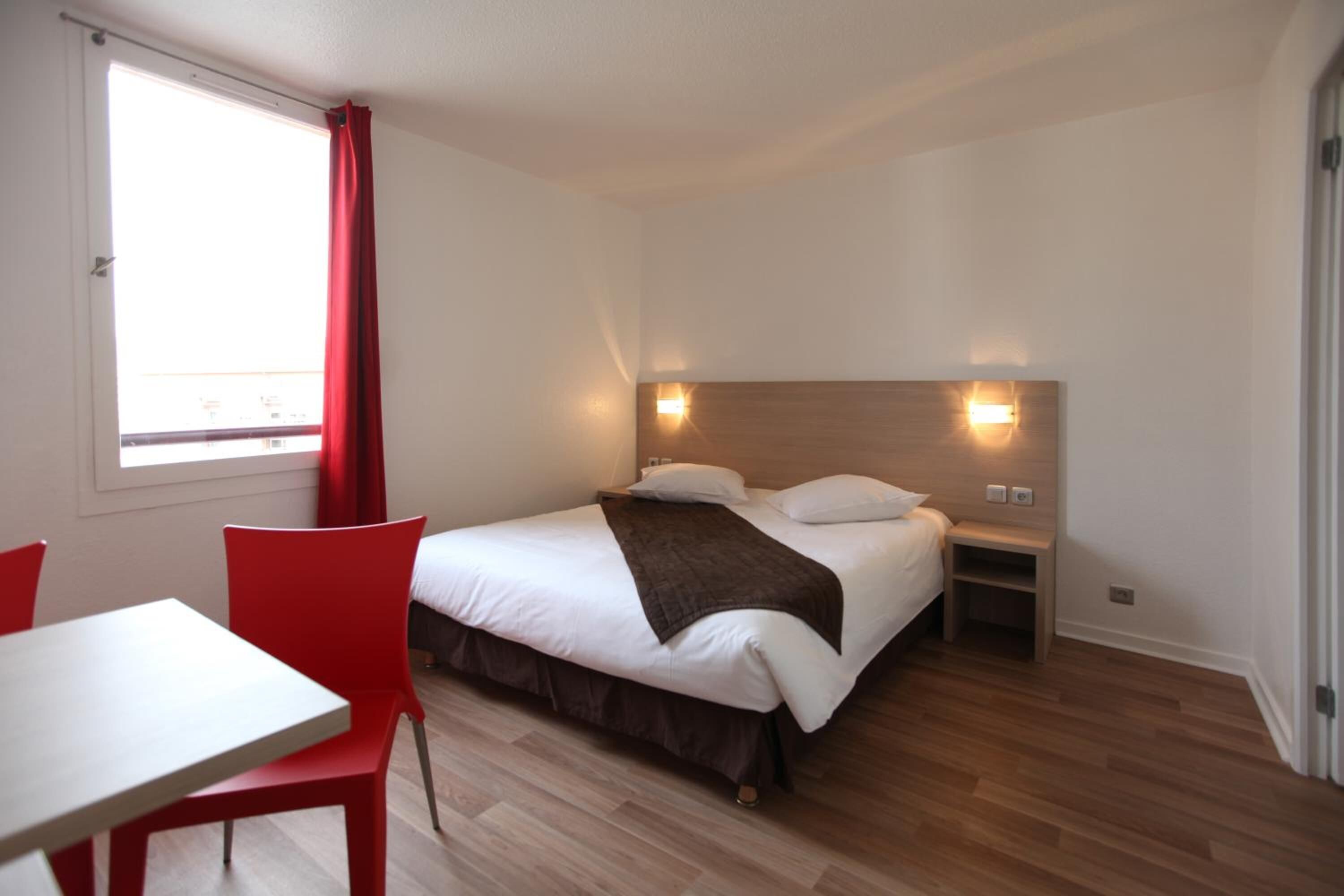 A hotel room with a bed, a bedside table, a nightstand, a window with red curtains, a white table, and a red chair.