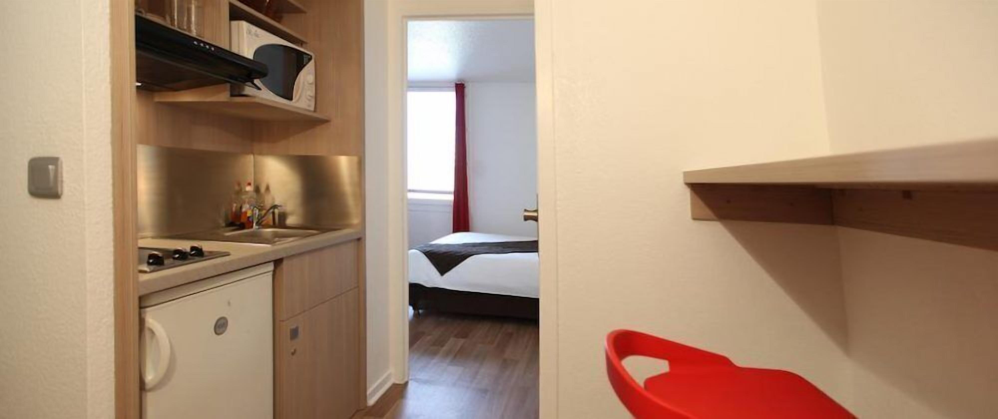 Cocina compacta con microondas, hornilla y pileta. Se ve una cama en la habitación contigua, y una silla roja al frente.
