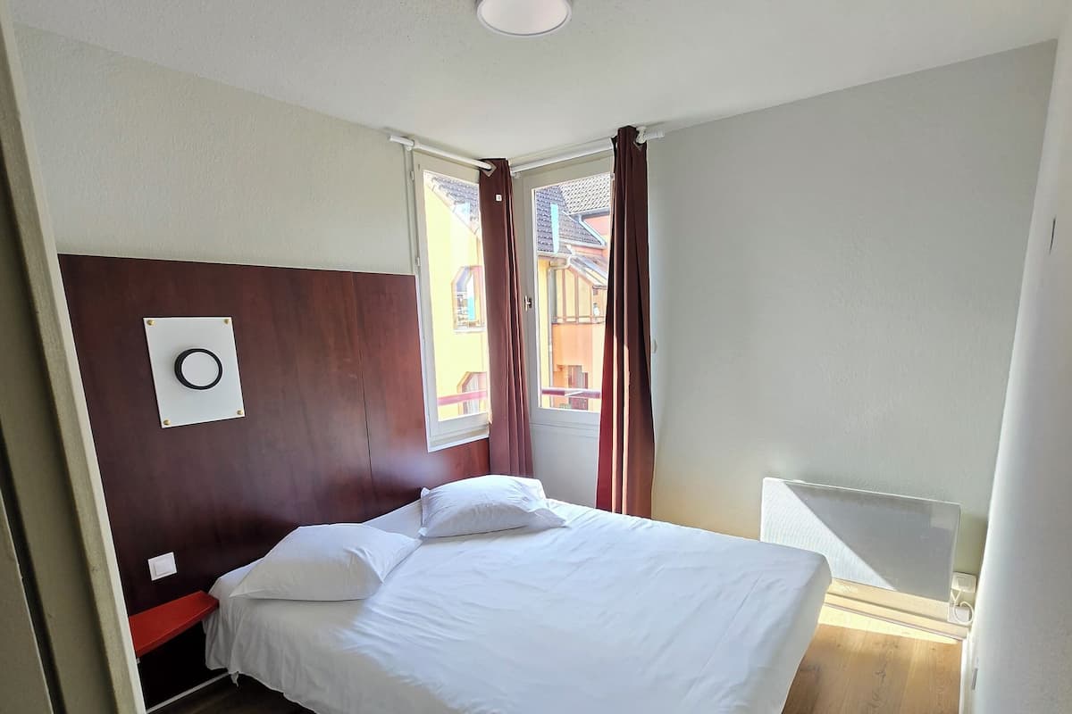A bedroom with a wooden floor, a bed with white bedding, a wooden headboard, a window with curtains, and a small bedside table.