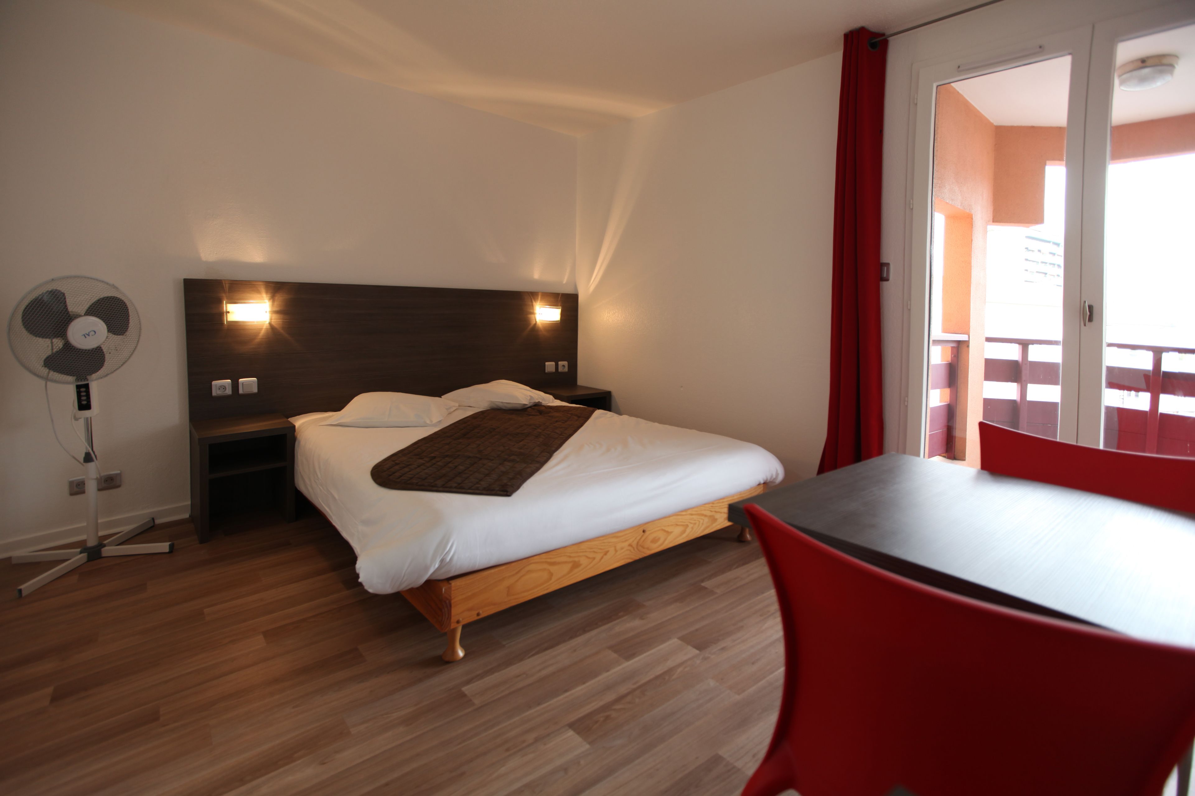 Une chambre avec un lit en bois, des tables de chevet, un ventilateur sur pied et une chaise rouge.