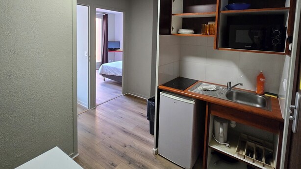 Eine kompakte Küche mit Kühlschrank, Spüle und Holz Arbeitsplatte. Ein Bett ist im angrenzenden Raum zu sehen.