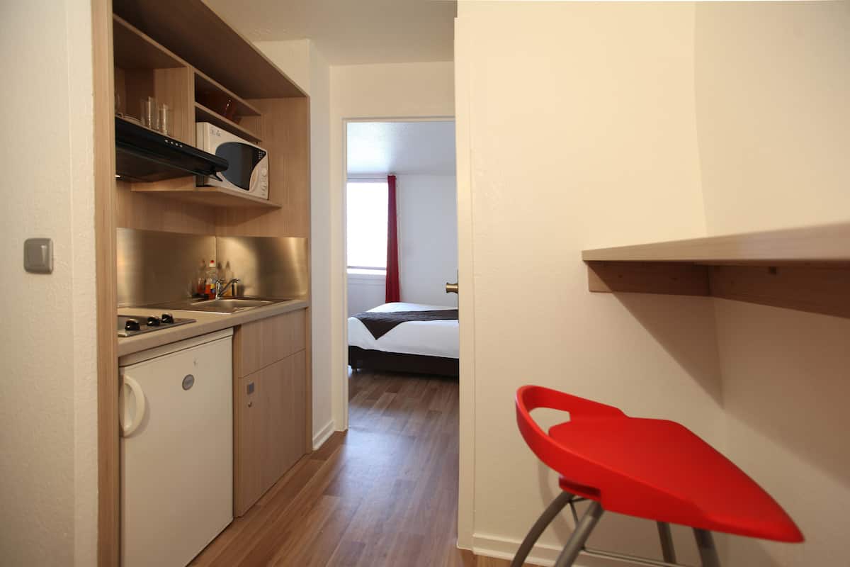 A compact kitchen with a microwave, sink, and cabinets, leading to a bedroom with a bed and window.