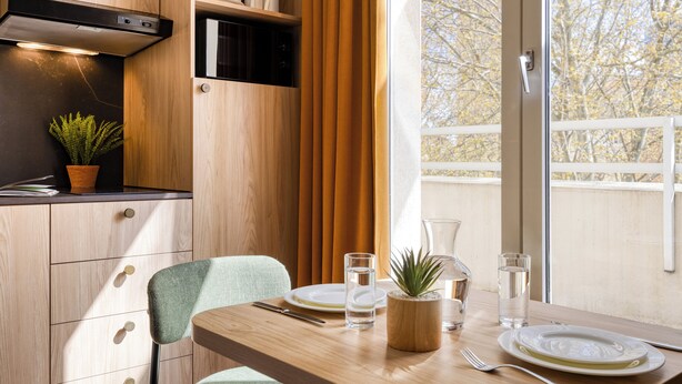 Una cocina moderna con armarios de madera, una mesa de comedor con sillas, una planta en maceta y vistas a árboles a través de una ventana.
