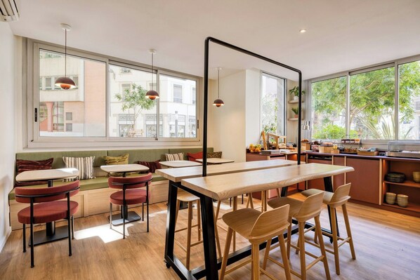 Un café moderno con una larga mesa de madera, sillas rojas y una zona de estar con un banco.