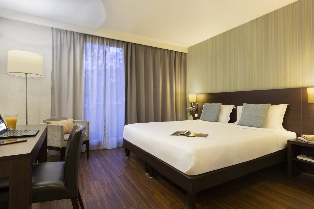 Une chambre d’hôtel avec un grand lit, un bureau avec un ordinateur portable, une chaise, une lampe et une fenêtre avec des rideaux.