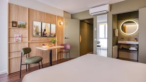 Habitación de hotel moderna con cama, una mesita con sillas, televisor montado en la pared, espejo con luz y baño visible a través de una puerta abierta.