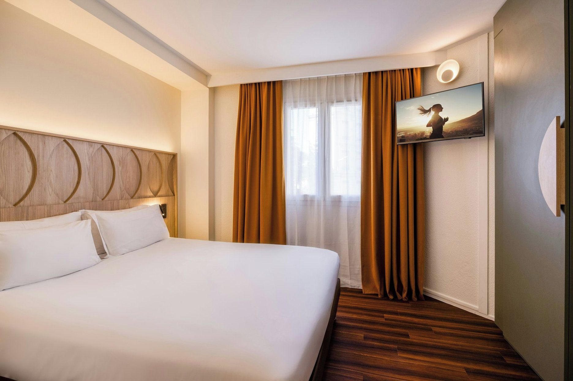 Hotellihuone, jossa on suuri sänky, seinälle asennettu televisio ja ikkuna, jossa on verhot.