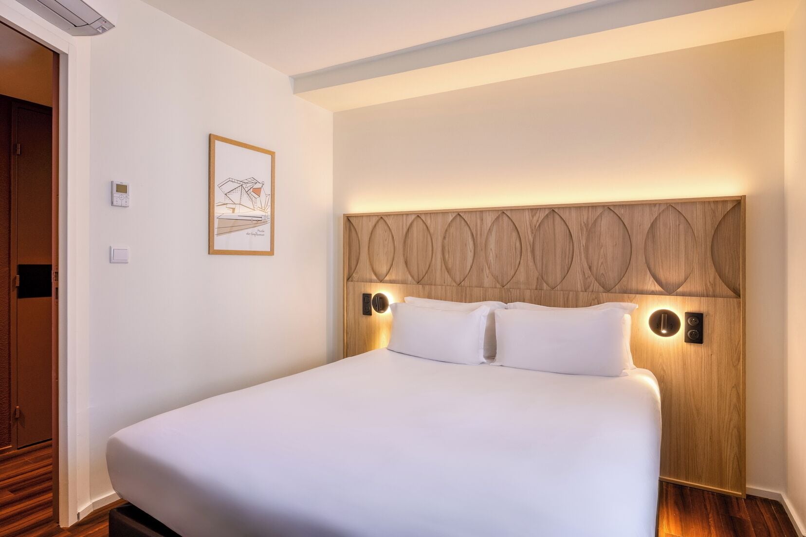 Ein Hotelzimmer mit einem großen Bett, einem hölzernen Kopfteil mit integrierter Beleuchtung, einem gerahmten Bild an der Wand und einer Tür links.