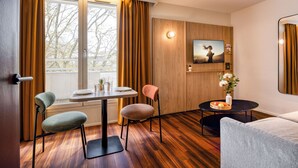 Una habitación moderna con piso de madera, una mesa de comedor con sillas, un sofá, un televisor montado en la pared y una pequeña mesa con un jarrón de flores.
