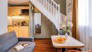 Un salón moderno con un sofá, una mesa de comedor con fruta, un florero con flores y una cocina al fondo.