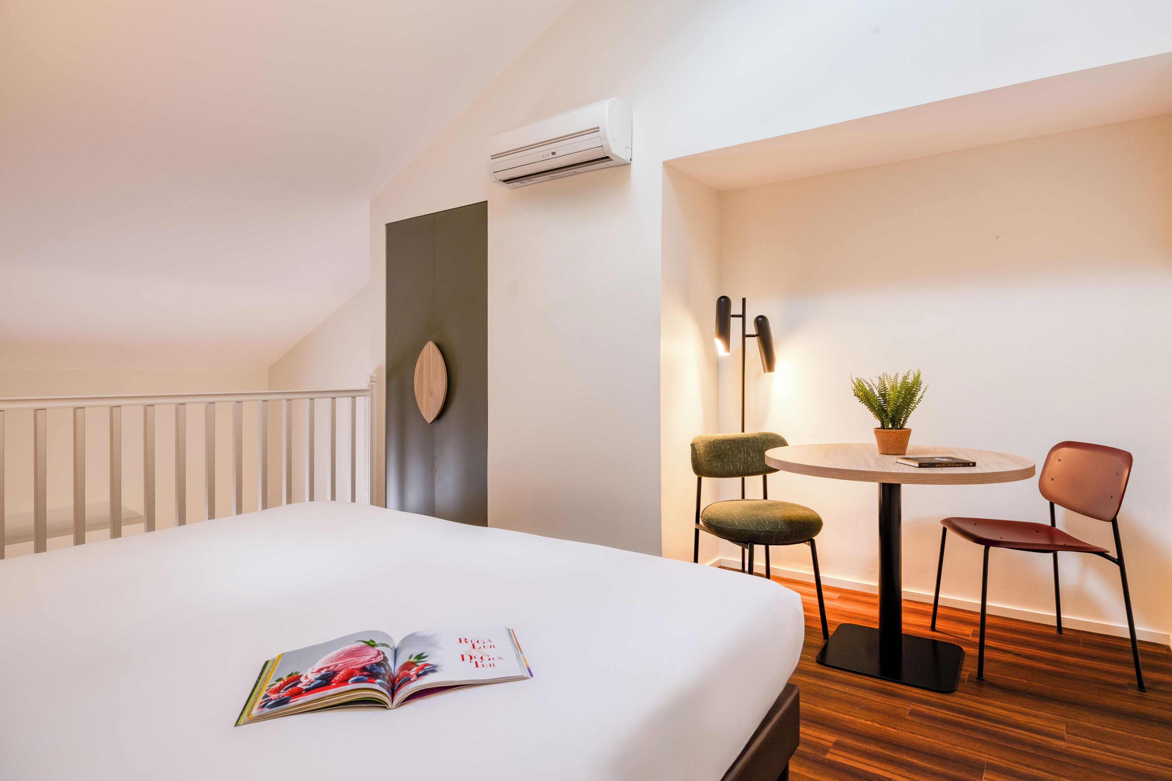 Una camera d'albergo con un letto, un tavolino con sedie, un condizionatore d'aria e un punto luce a parete.