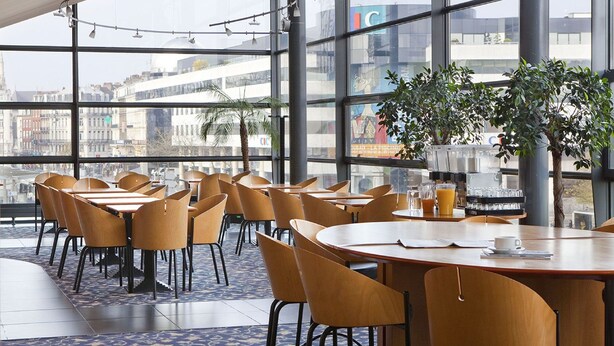 Een moderne eetruimte met ronde tafels en stoelen, grote ramen met uitzicht op de stad, en een koffieapparaat op de tafels.