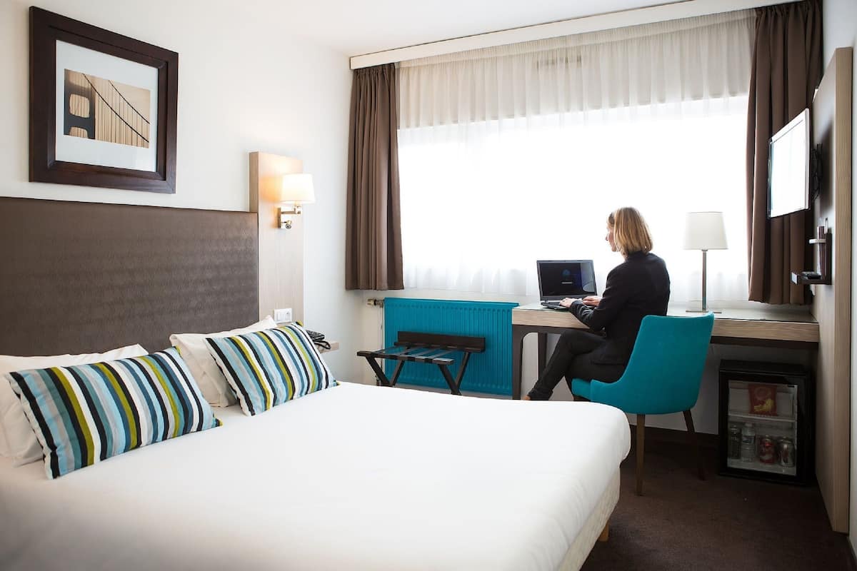 A hotel room with a bed, a desk, a chair, a computer, a window with curtains, a framed picture on the wall, and a minibar.