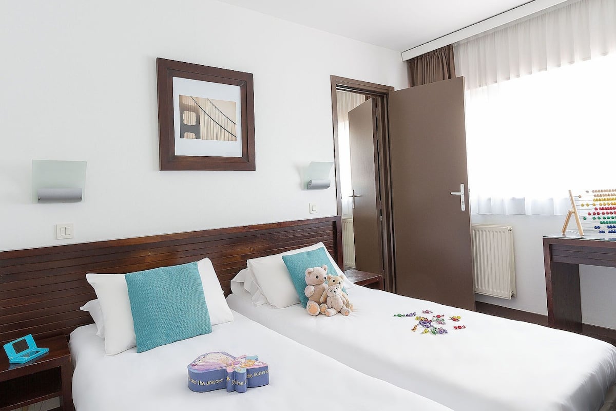 A hotel room with a bed, a nightstand, a desk with a lamp, a framed picture on the wall, and a window with curtains.
