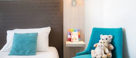Une chambre d’hôtel avec un lit, un fauteuil turquoise, une table de chevet avec des livres et une lampe fixée au mur.