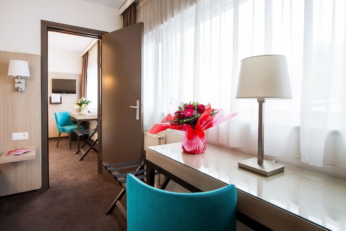A hotel room with a desk, a chair, a lamp, a bouquet of flowers, and a television on the wall.
