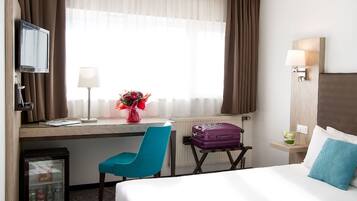 Habitación de hotel con cama, escritorio con computadora, silla, maleta, refrigerador pequeño y ventana con cortinas.