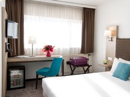 Habitación de hotel con cama, escritorio con computadora, silla, maleta, refrigerador pequeño y ventana con cortinas.