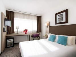 Habitación de hotel con cama, escritorio, silla, televisor y ventana con cortinas.