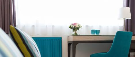 A hotel room with a bed, a desk with a chair, a lamp, and a window with curtains.