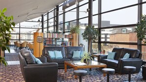 Een ruime zithoek met leren banken, een salontafel en barstoelen. Boekenkasten vol boeken en potplanten zorgen voor een gezellige sfeer. Grote ramen bieden uitzicht op de stads skyline.