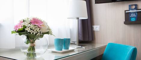 Een hotelkamer met een glazen tafel, een blauwgroene stoel, een vaas met bloemen, twee blauwe kopjes en een lamp.