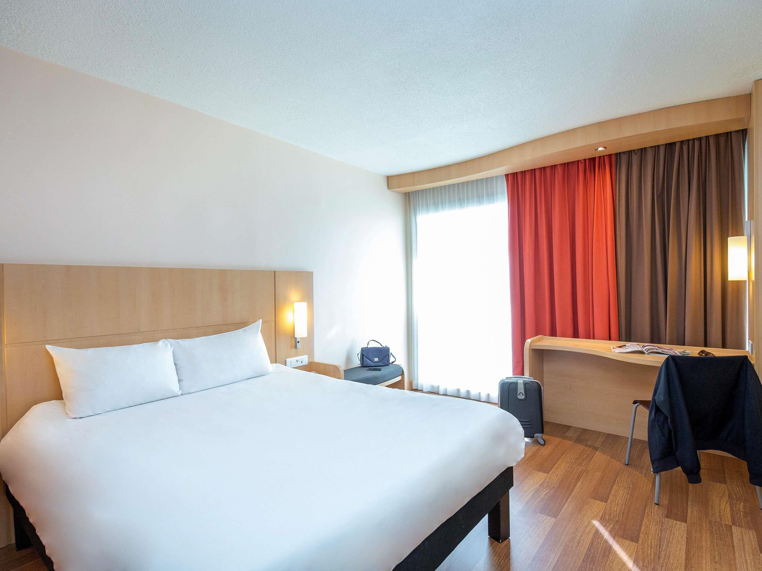 Habitación de hotel con una cama grande, un escritorio con silla, una maleta y una ventana con cortinas.
