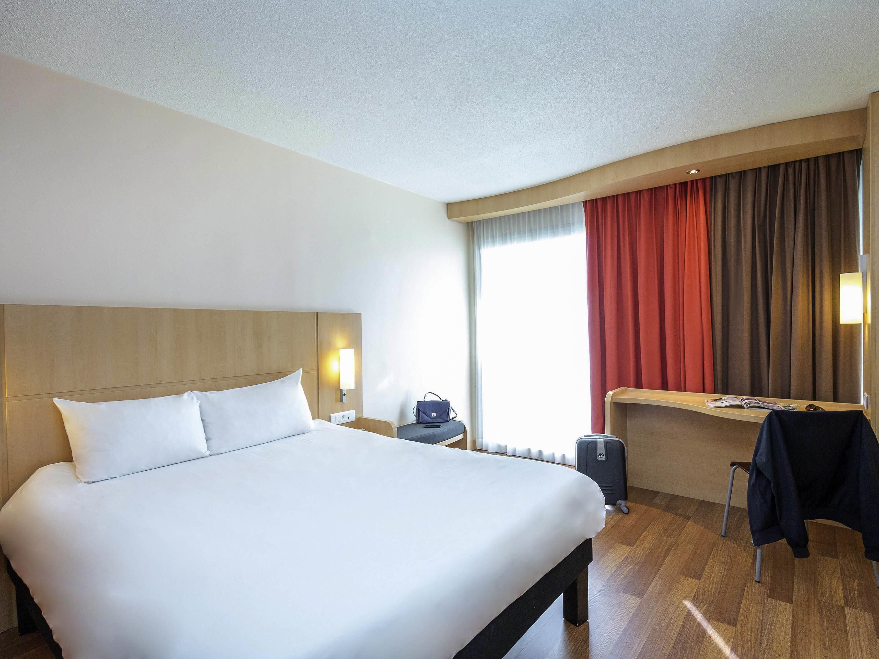 A hotel room with a large bed, a desk, a chair, a suitcase, and a window with curtains.