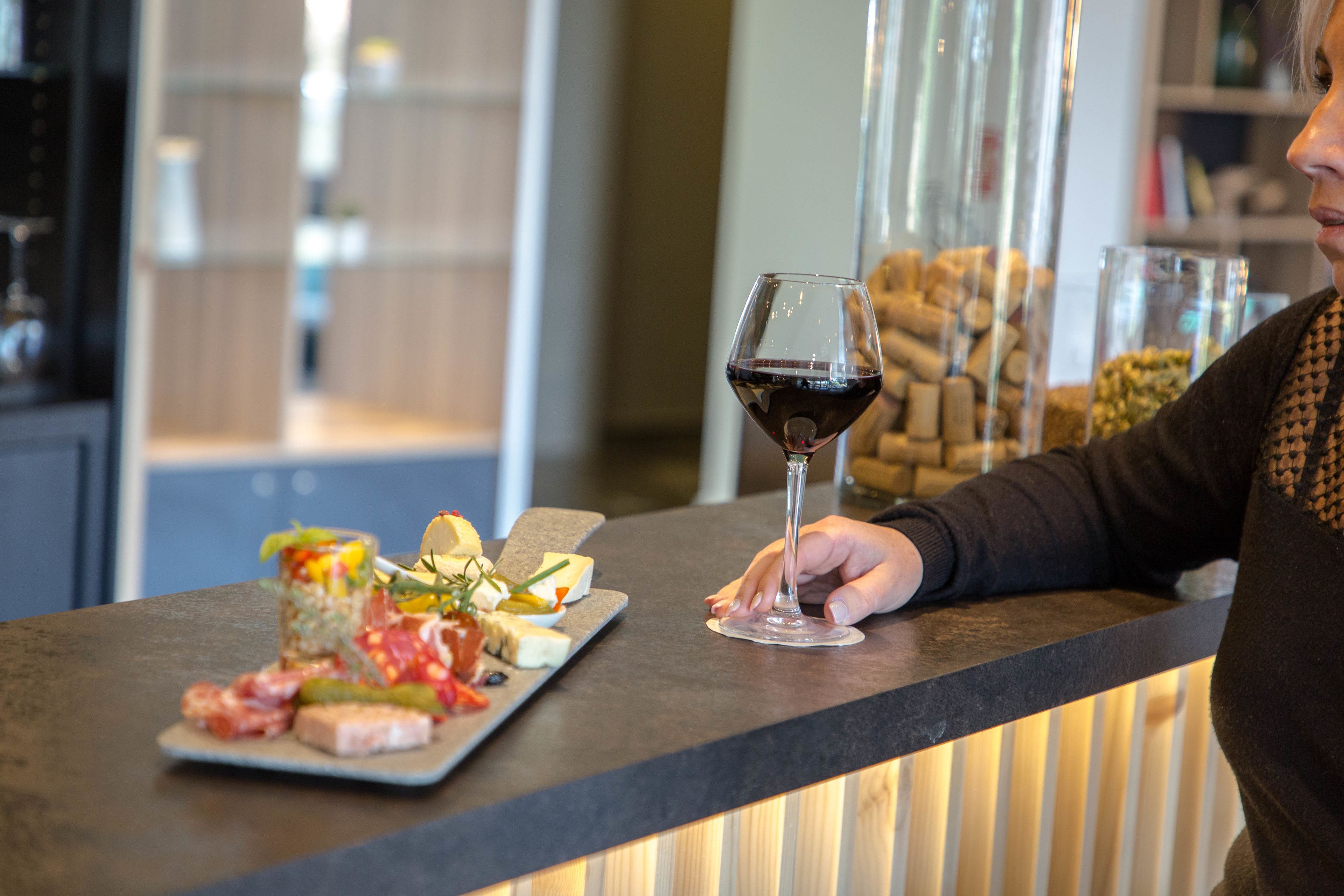 A bar area with a glass of red wine, a cheese board with various cheeses and meats, and a bottle of wine behind the bar.