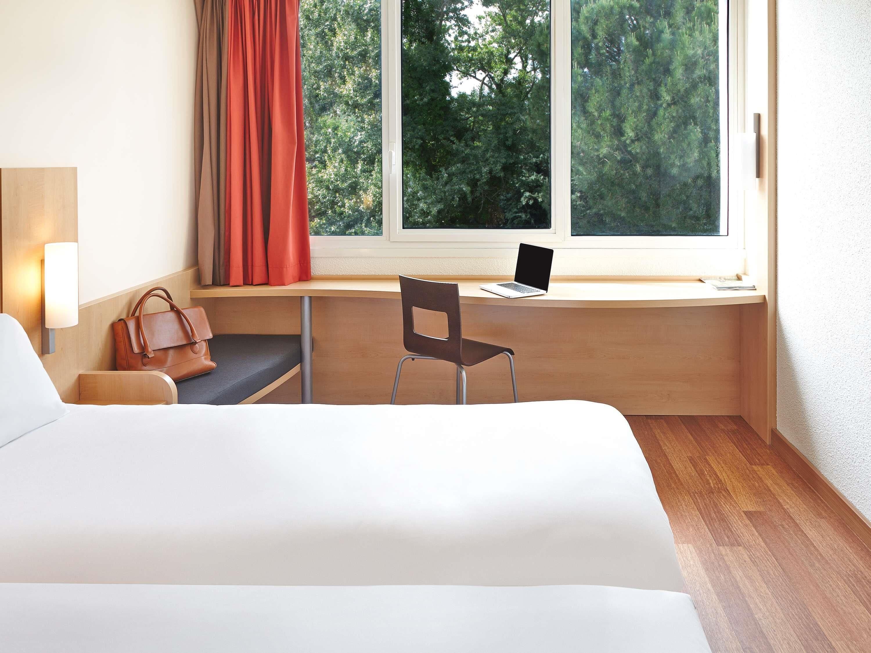 A hotel room with a bed, a desk with a laptop, a chair, a handbag, and a window with curtains.