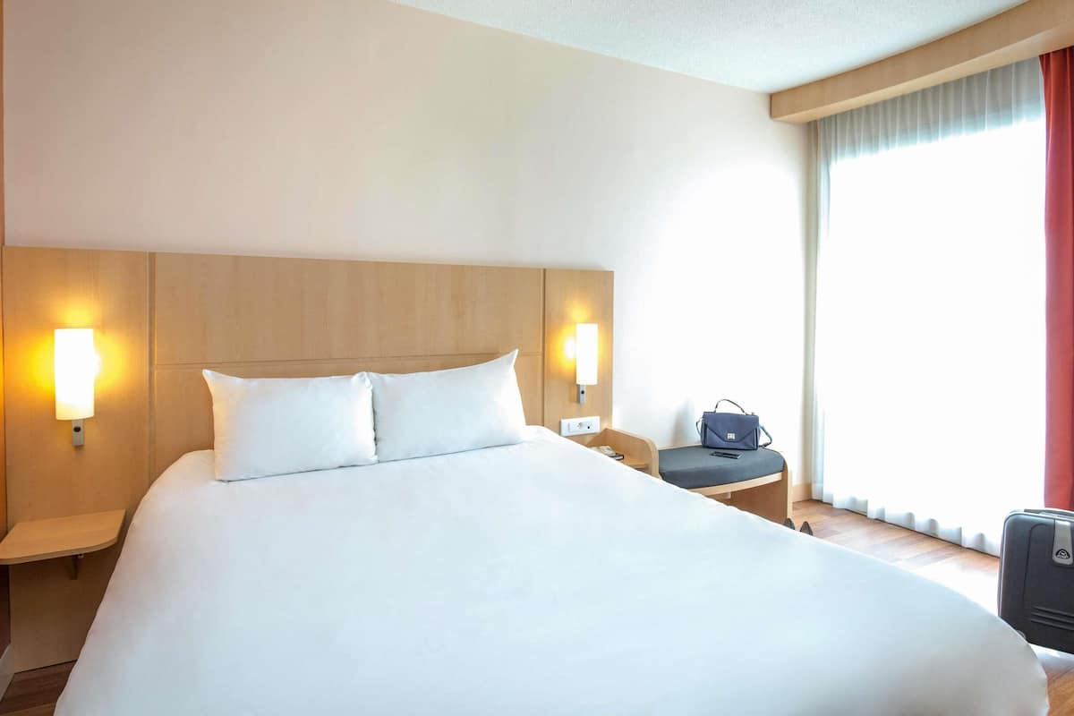 Habitación de hotel con una cama grande, dos lámparas de noche, una silla, un escritorio y una maleta.