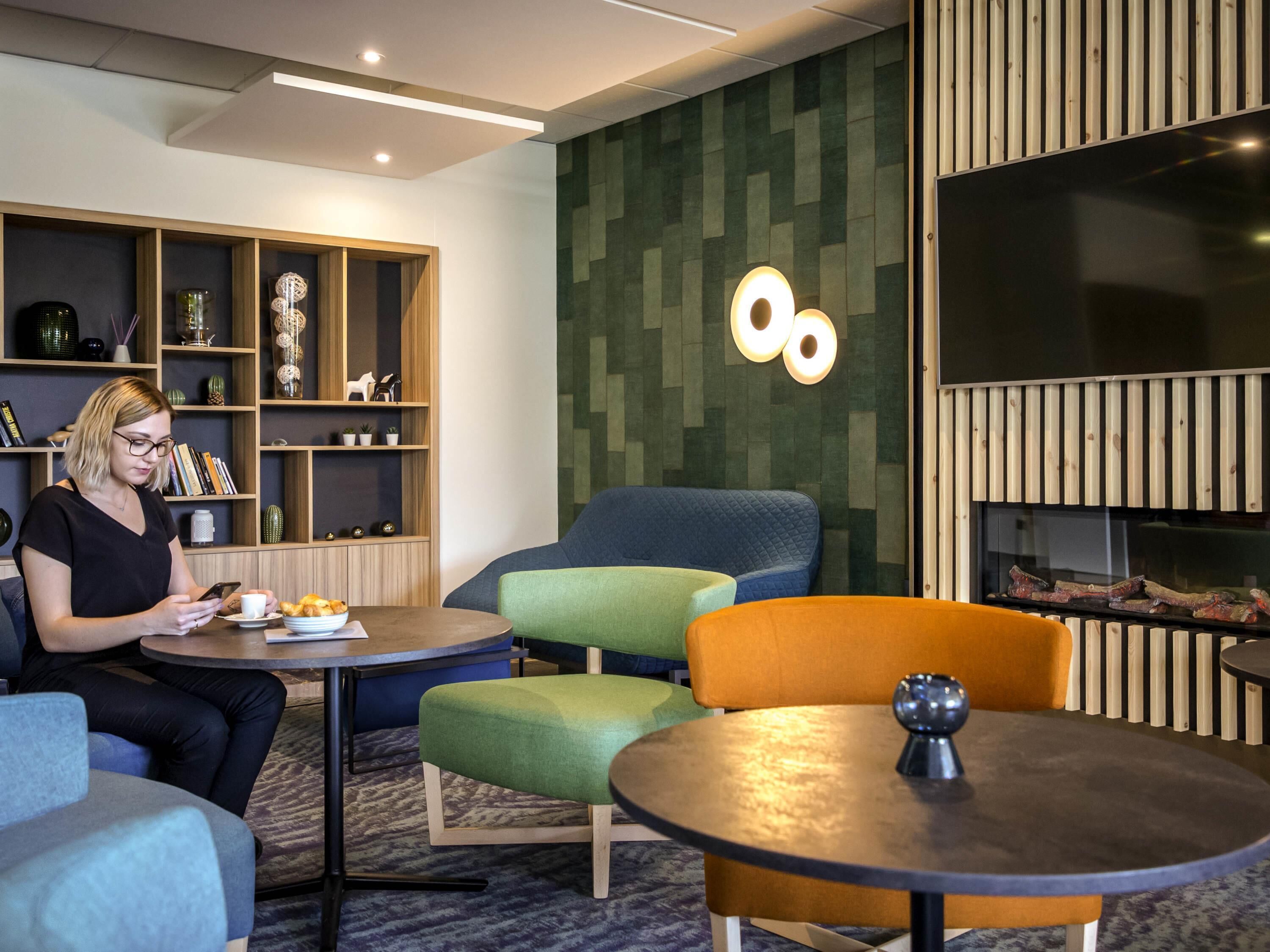 A modern lounge area with a seating arrangement including a sofa, chairs, and a round table. There are shelves with decorative items and a wall-mounted light fixture.