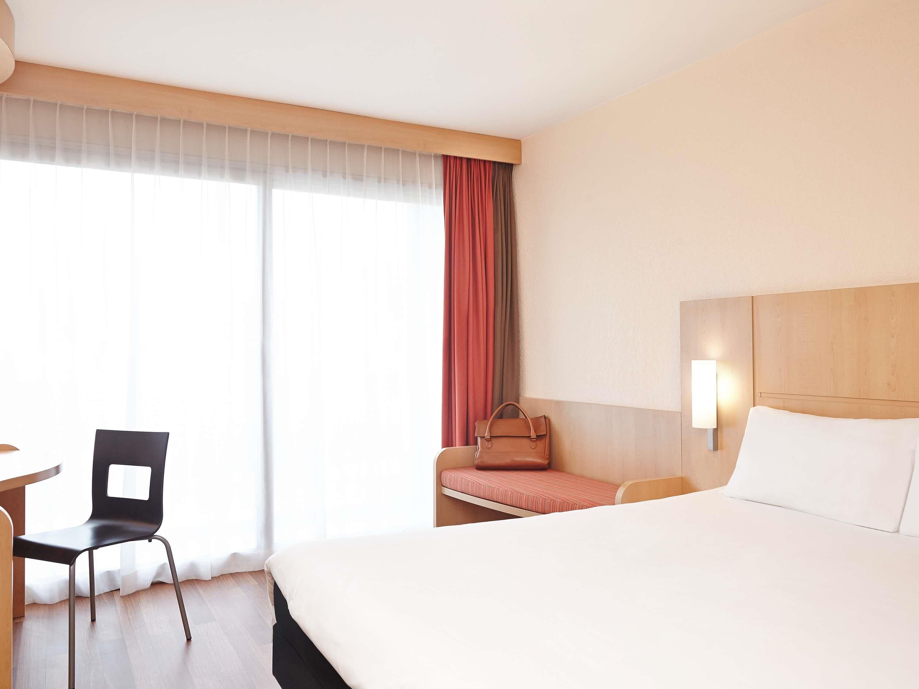 A hotel room with a bed, a desk, a chair, a window with curtains, and a wall-mounted lamp.