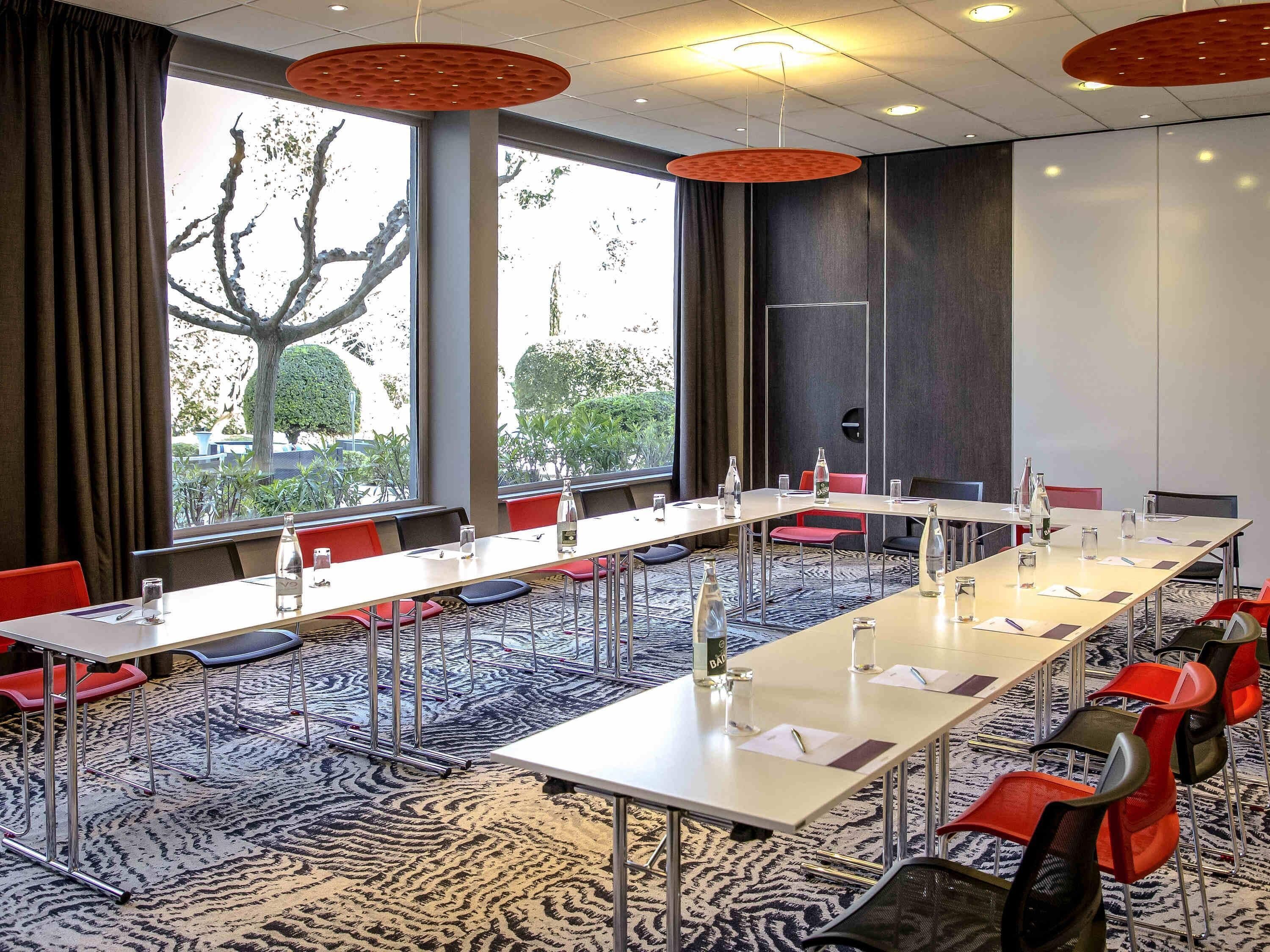 A conference room with long tables, red chairs, and large windows with a view of trees.