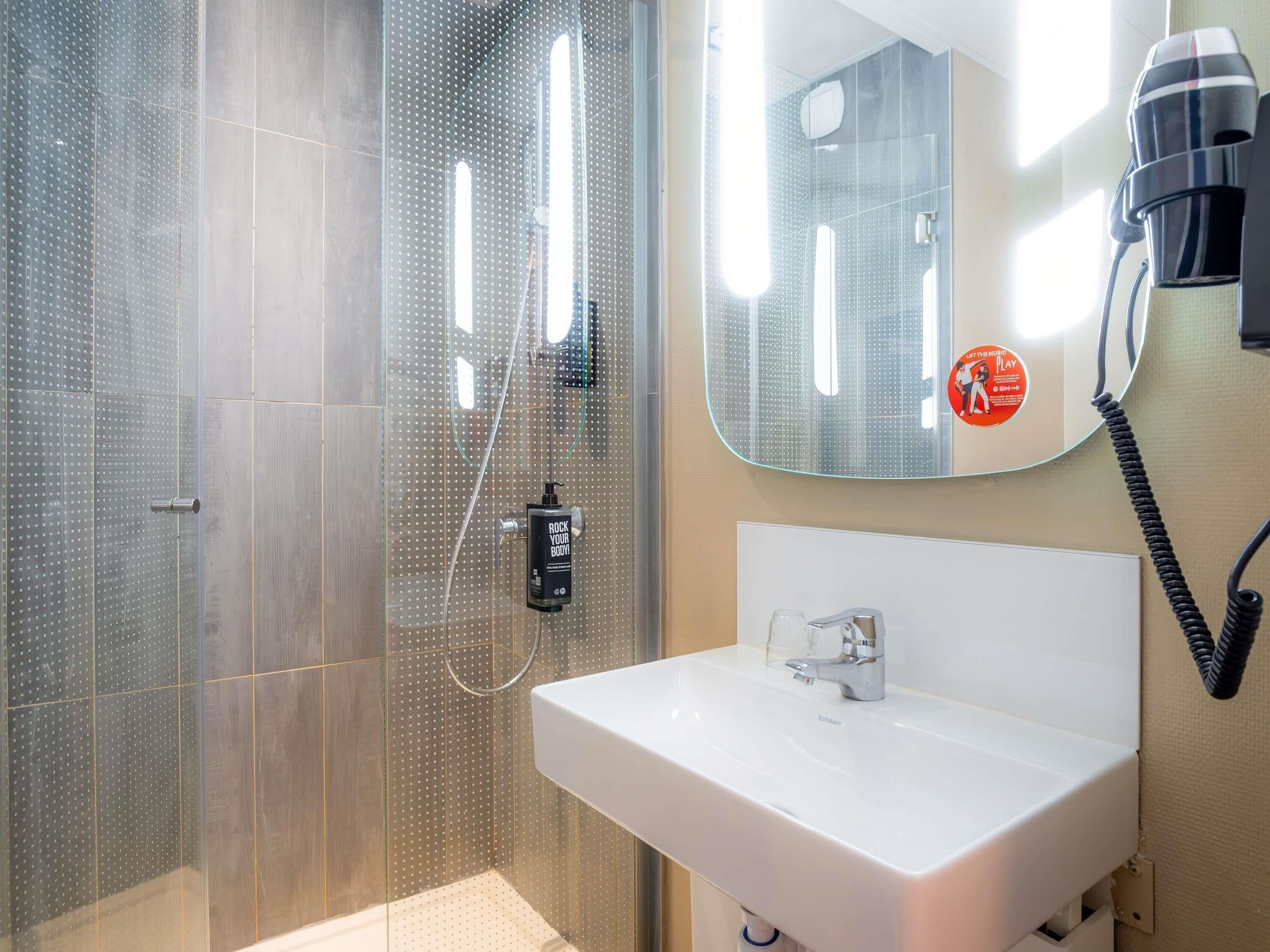 Un baño moderno con cabina de ducha de vidrio, un lavamanos blanco y un espejo con secador de pelo incorporado.