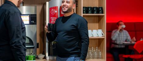 Un hombre sostiene una taza negra, de pie frente a una máquina de café y una estantería con tazas.