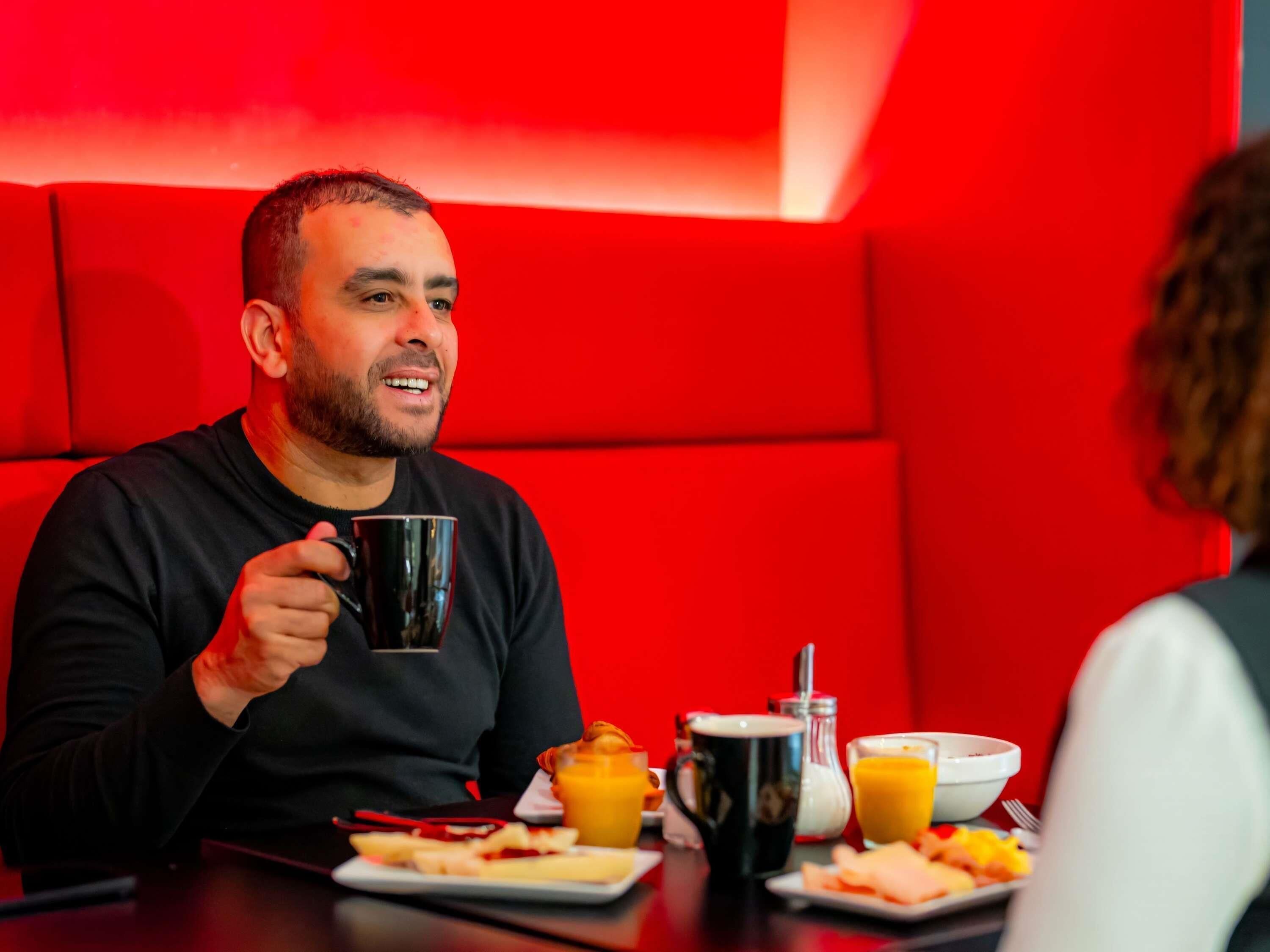 Eine Person sitzt an einem Tisch mit einer schwarzen Tasse, einem Teller mit Essen und einem Glas Orangensaft.