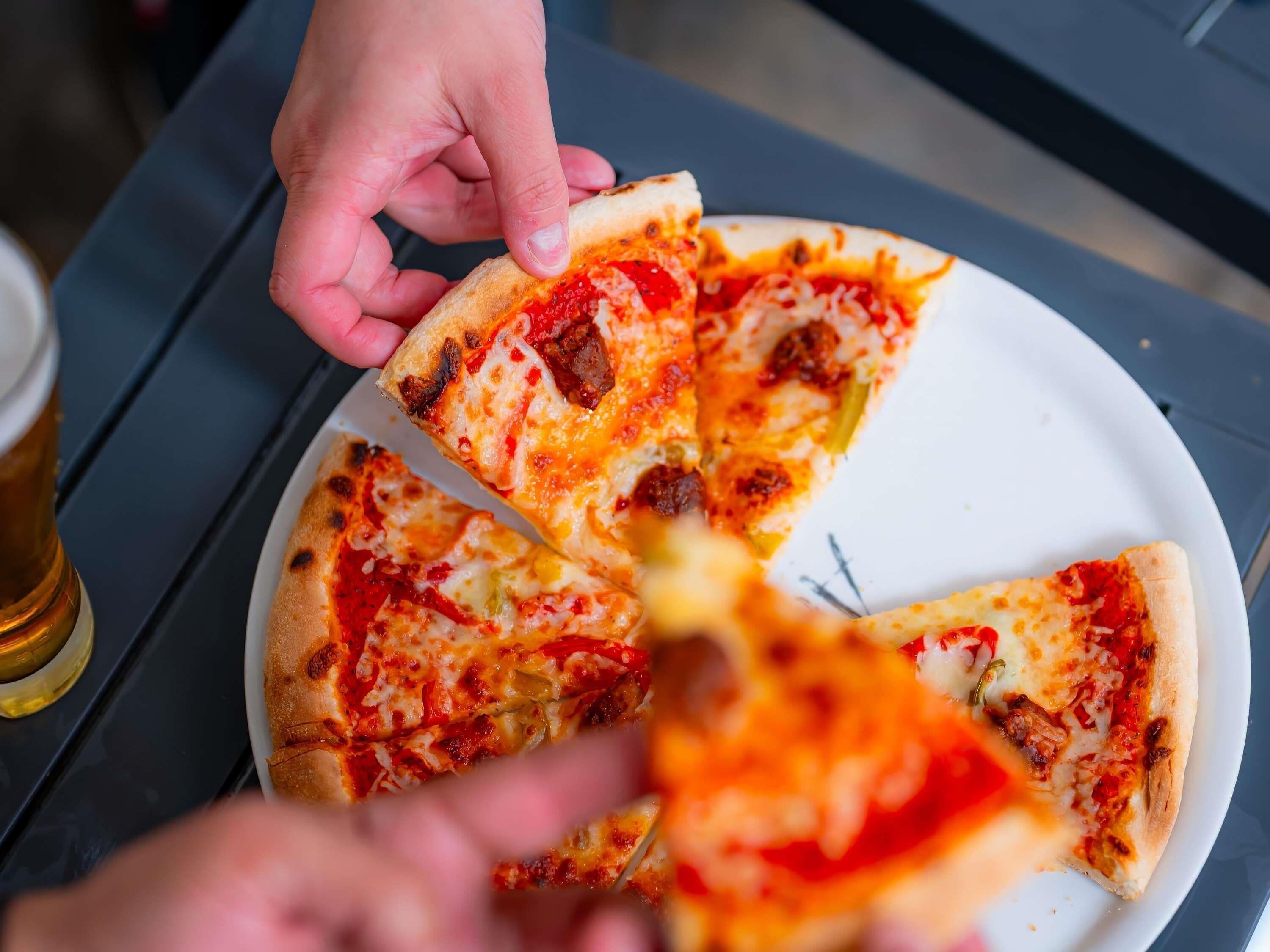A hand holding a slice of pizza with visible toppings like cheese, pepperoni, and possibly vegetables, on a white plate.