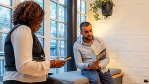 Two people sitting on a bench in a room with large windows and a hanging plant. - ibis Paris Ornano Montmartre Nord 18ème (Paris)