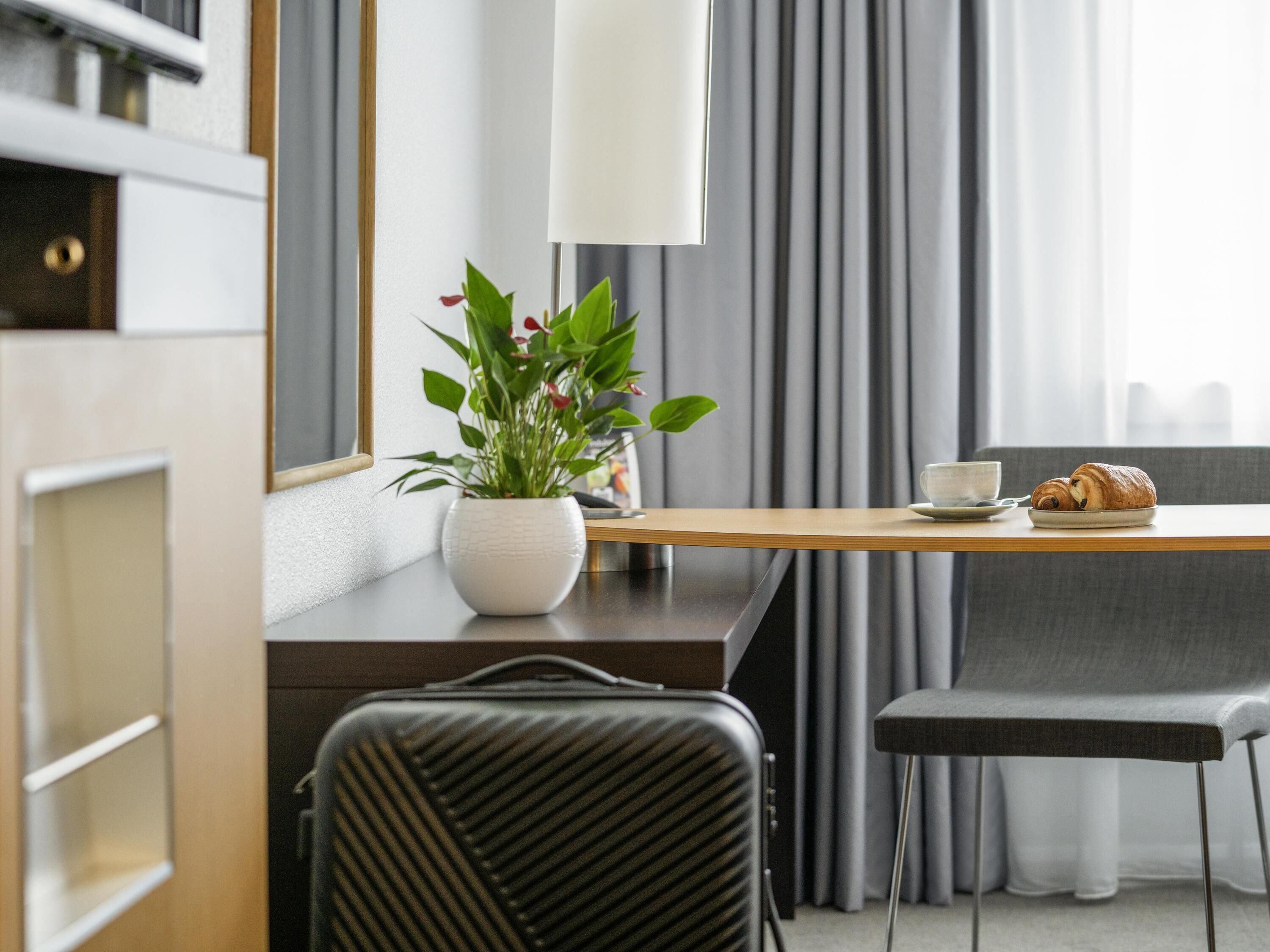 Una habitación de hotel moderna con un escritorio de madera, una planta en maceta, una silla, una cama y una ventana con cortinas.