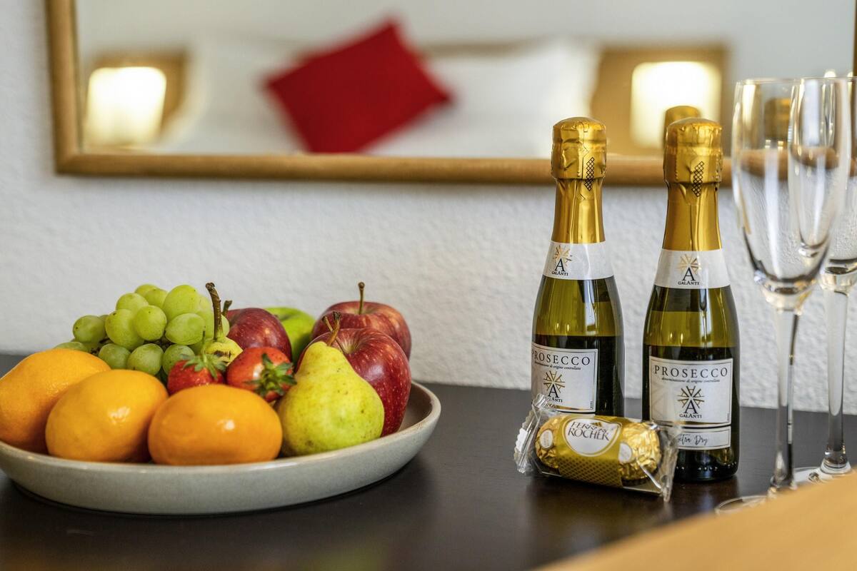 Una mesa con un bol de fruta, dos botellas de Prosecco, un bombón envuelto y dos vasos.